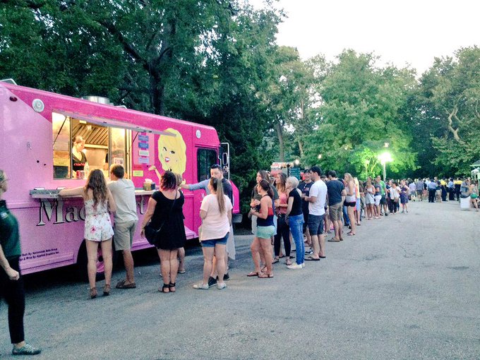 People standing in line outside a pink food truck with a cartoon character, in a park with trees.