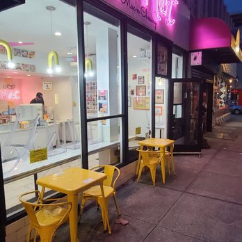 Yellow tables and chairs outside a cafe with large glass windows, interior visible, pink neon sign above, sidewalk scene.