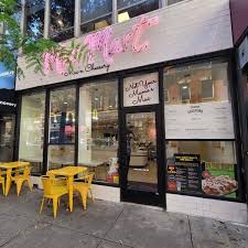 Cafe storefront with large glass windows, yellow outdoor tables and chairs, and a sign with pink lettering. Confidence 0.9