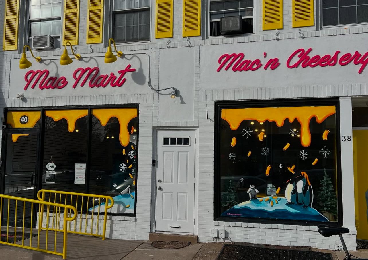 Storefront with yellow window frames, signs reading 'Mac Mart' and 'Mac'n Cheese,' and large display windows with penguin illustrations.