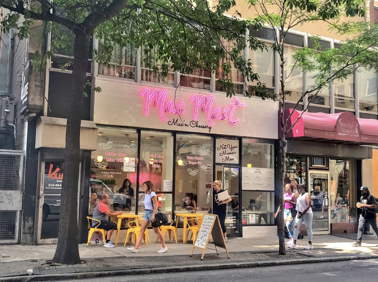 People sit at yellow tables outside a cafe with pink signage, trees line the sidewalk, and pedestrians walk by.