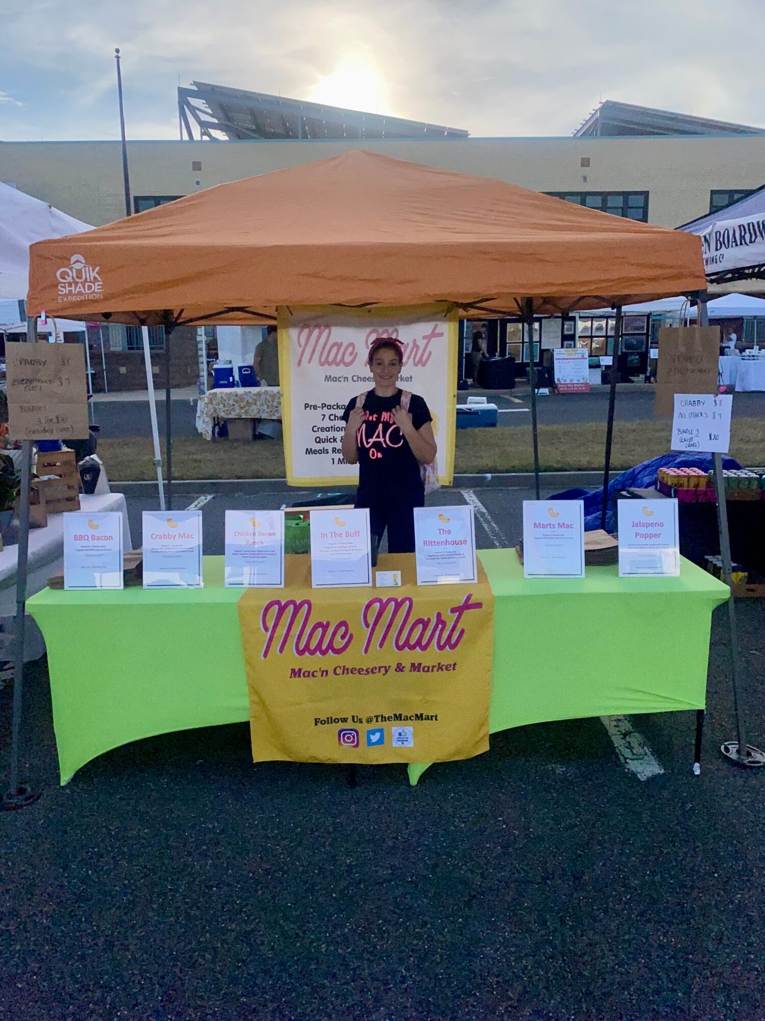 Person standing behind a table with signs and a yellow banner, under an orange canopy at an outdoor event.
