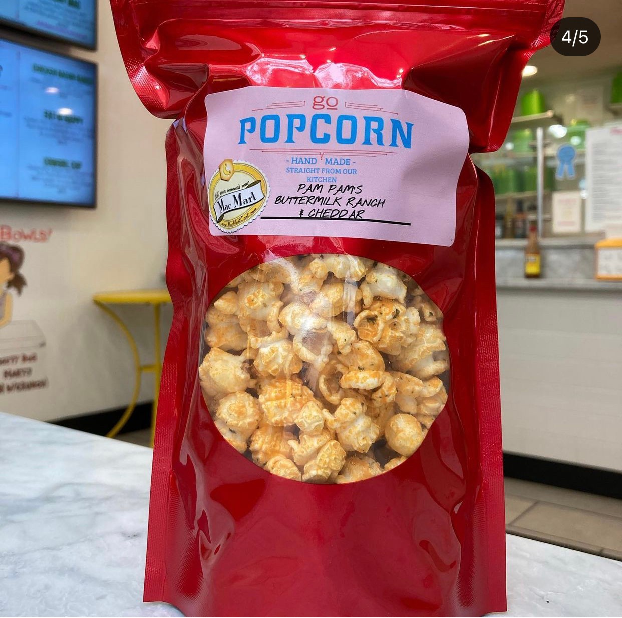 Red bag of popcorn with a label, transparent window showing popcorn inside, in a store setting.