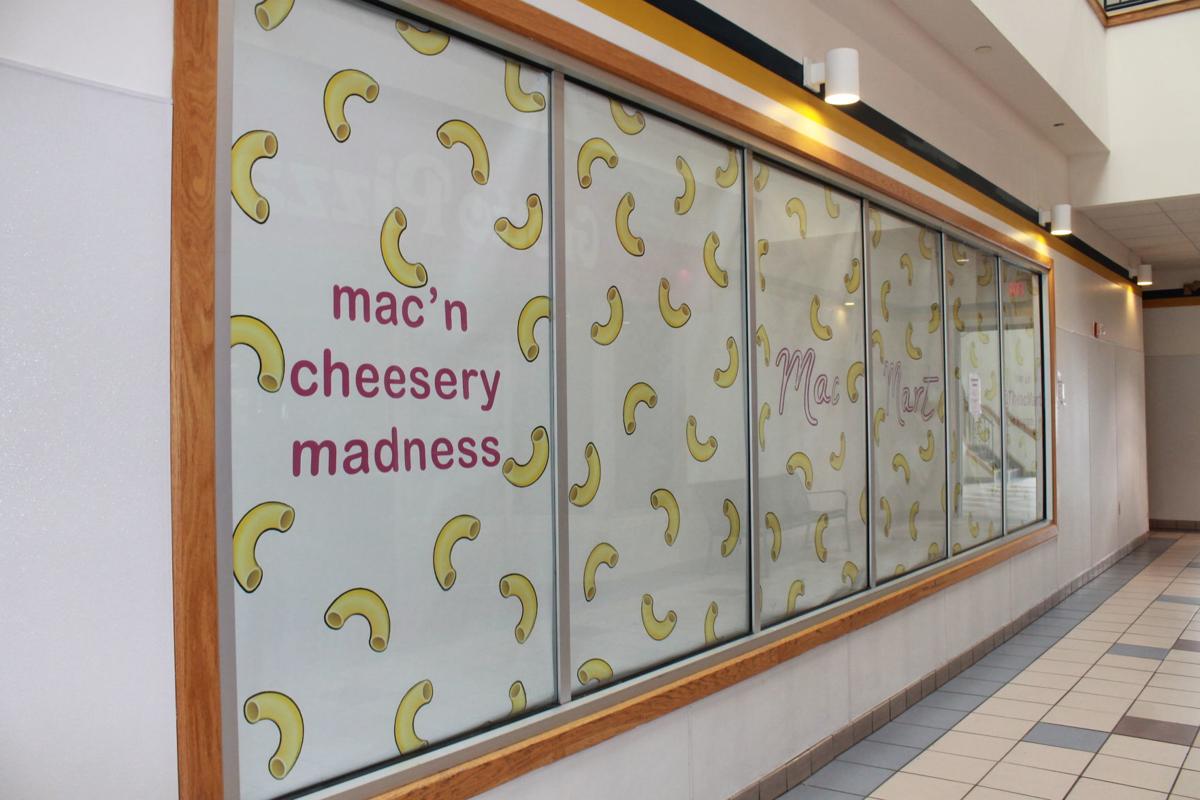 Store window with banana pattern and pink text reading 'mac'n cheesery madness' on glass, tiled floor, and ceiling lights.