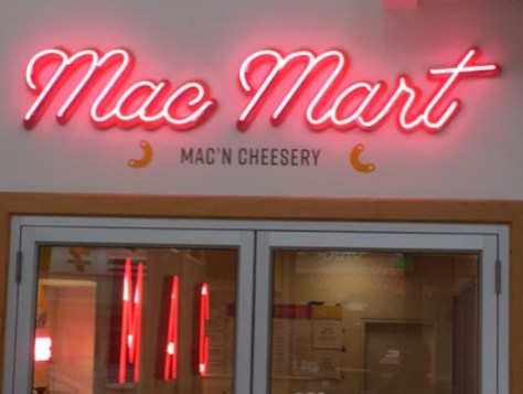 Neon sign reads 'Mac Mart' with smaller text 'MAC'N CHEESERY' below, above a glass door.