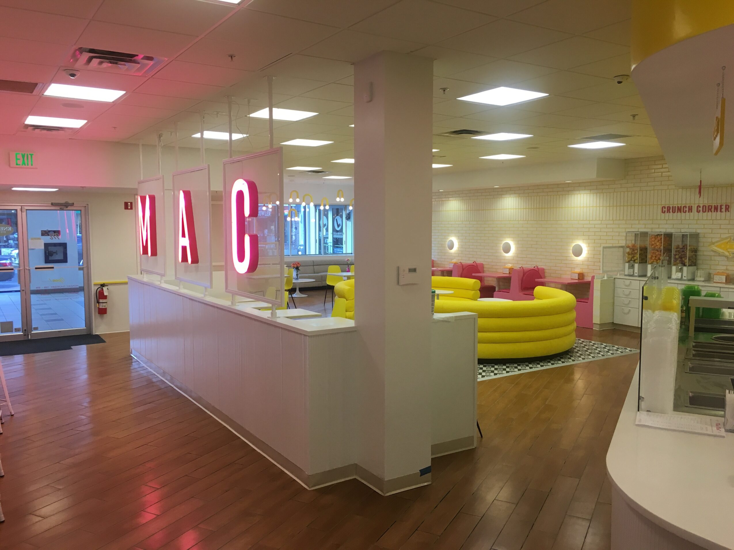 Interior of a store with pink illuminated letters spelling 'MAC', seating area with yellow and pink couches, and a wooden floor.