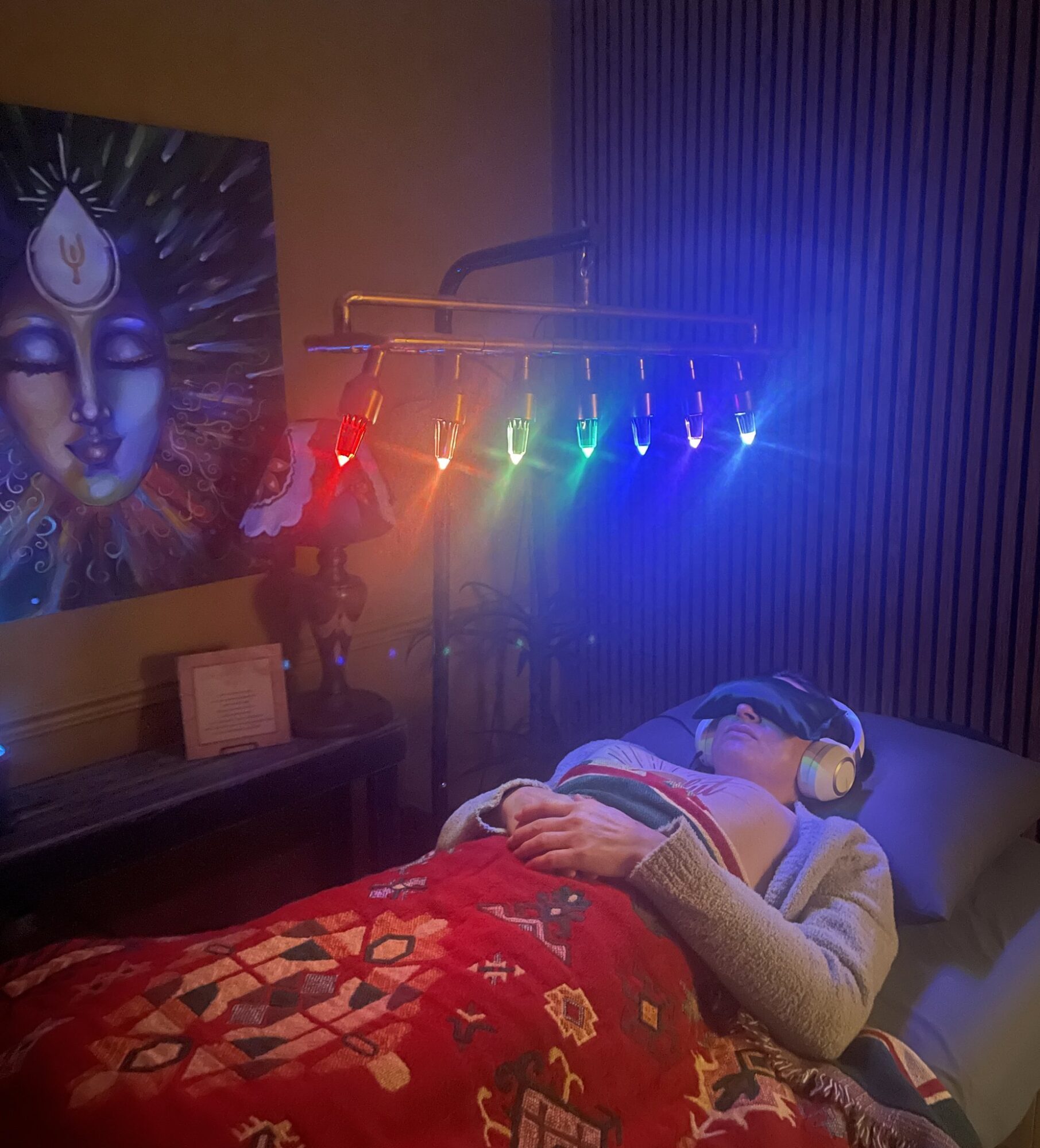 Person lying on bed with headphones, colorful lights above, artwork on wall, and a framed picture on a table.