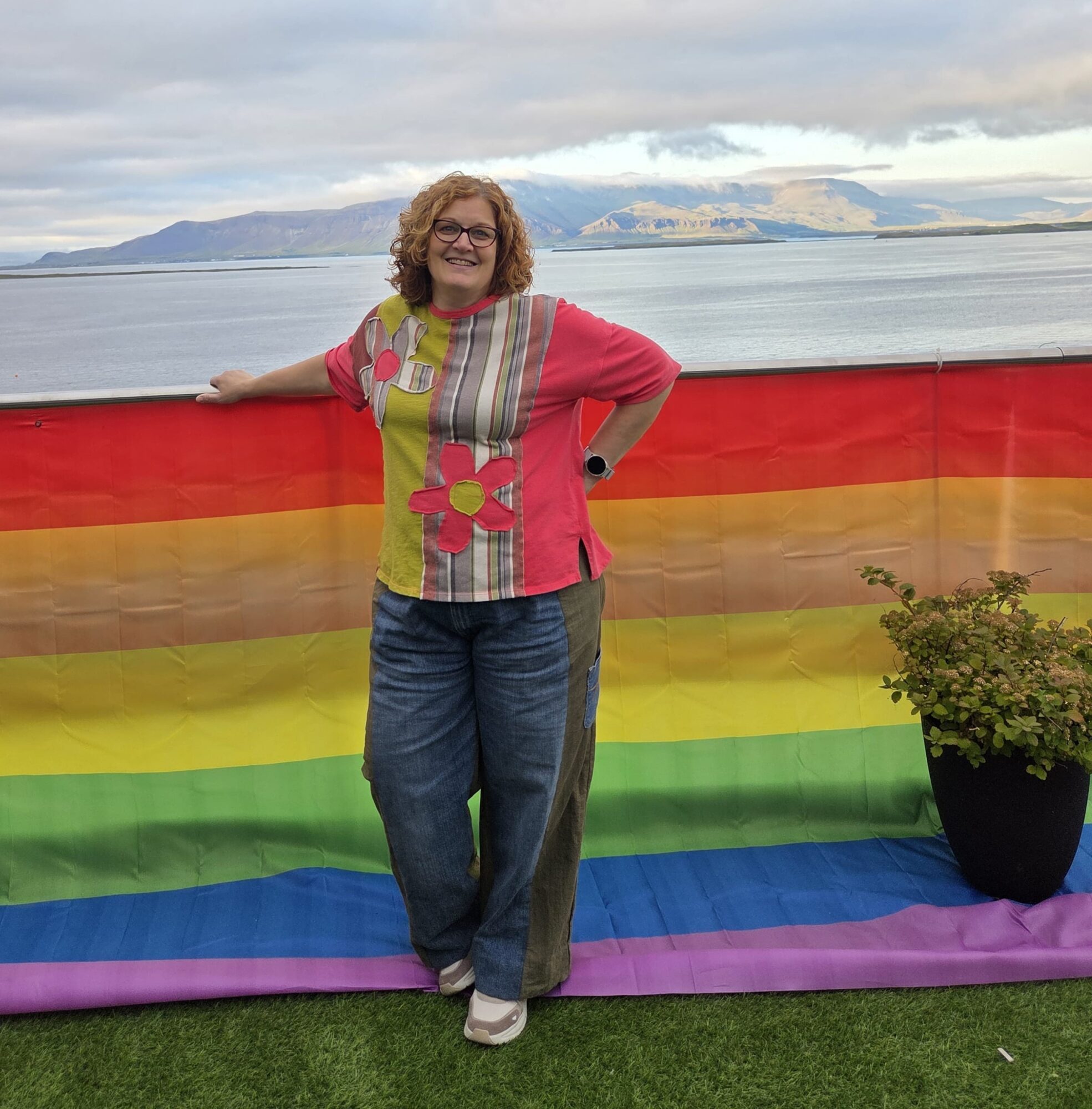 Woman standing outdoors in front of a rainbow flag, with a body of water and mountains in the background.