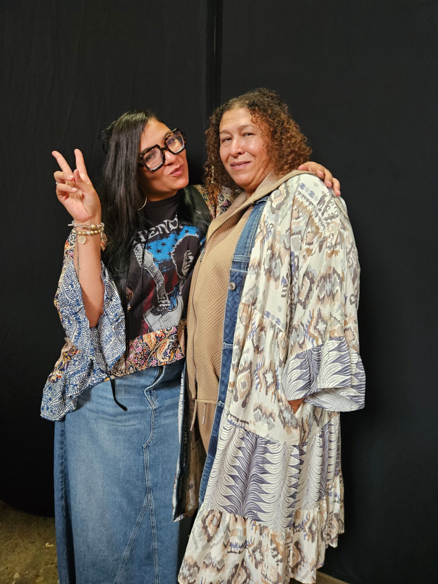 Two women standing, one with curly hair and the other with straight hair and glasses, posing together. The woman with curly hair is lying down on her side.