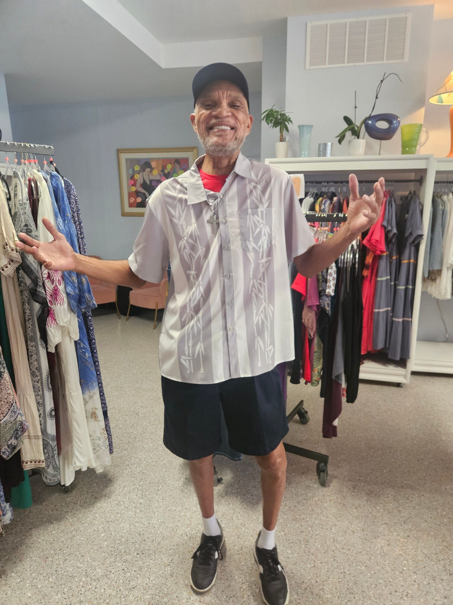 Person standing in a room with clothing racks, smiling, wearing a white shirt, black shorts, and a cap.