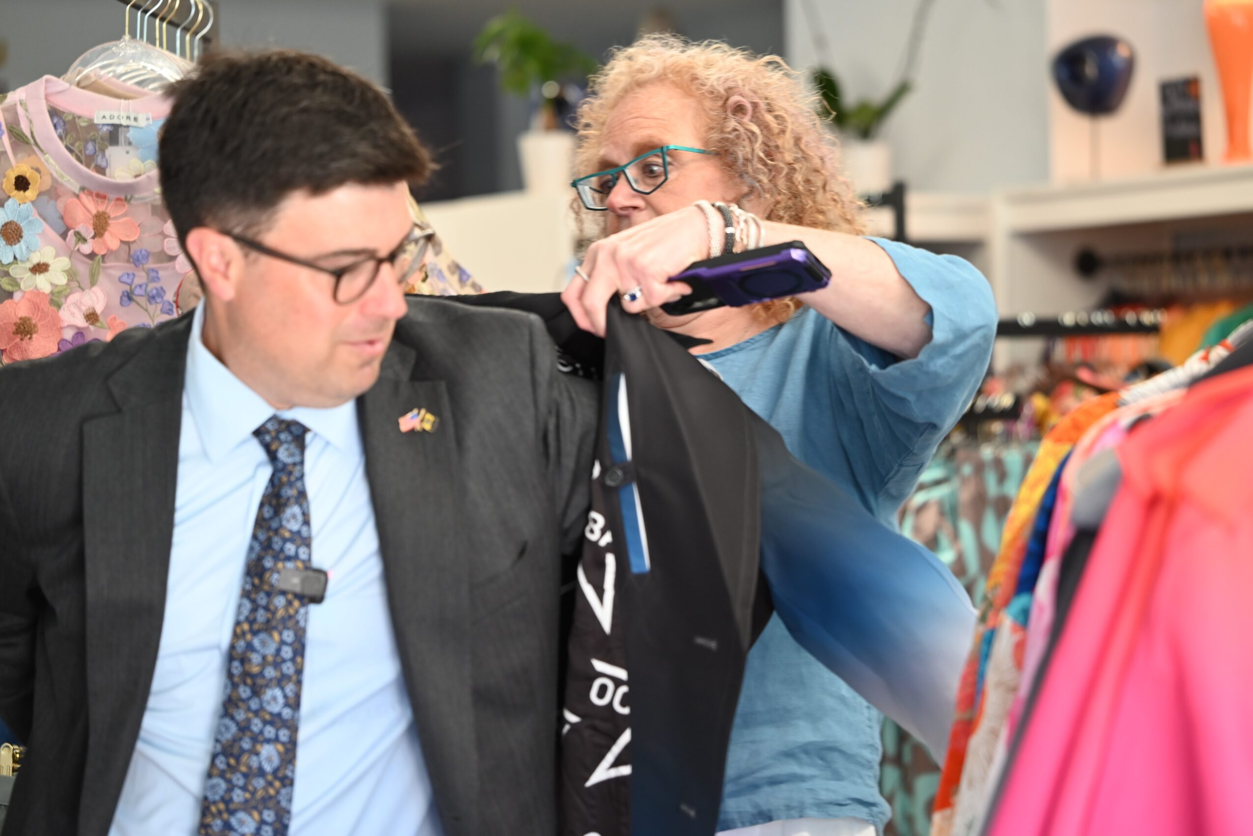 A woman helps a man try on a black jacket in a clothing store, with colorful clothes in the background.