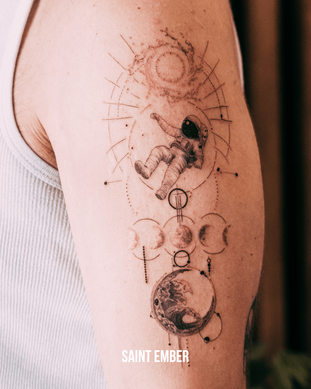Tattoo of an astronaut floating above a planet with geometric and celestial designs, labeled 'SAINT EMBER'.