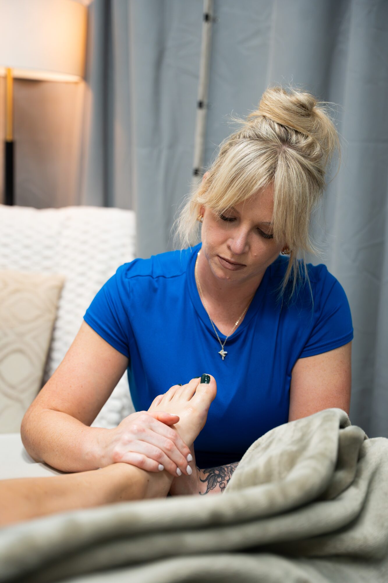 Woman with blonde hair in a bun wearing a blue shirt, sitting indoors, holding a person's hand, with a bed and curtains in background.