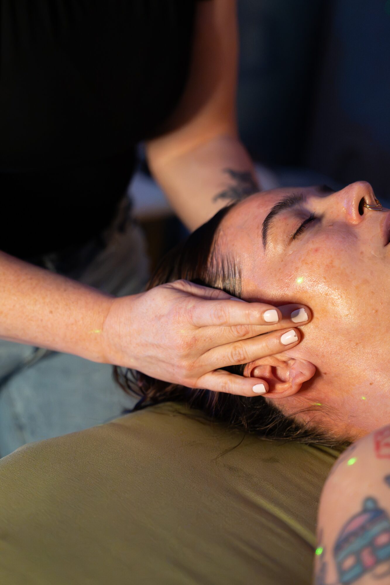 Person receiving a facial massage with eyes closed, lying on a surface, with hands gently on face, tattoos visible on arm.