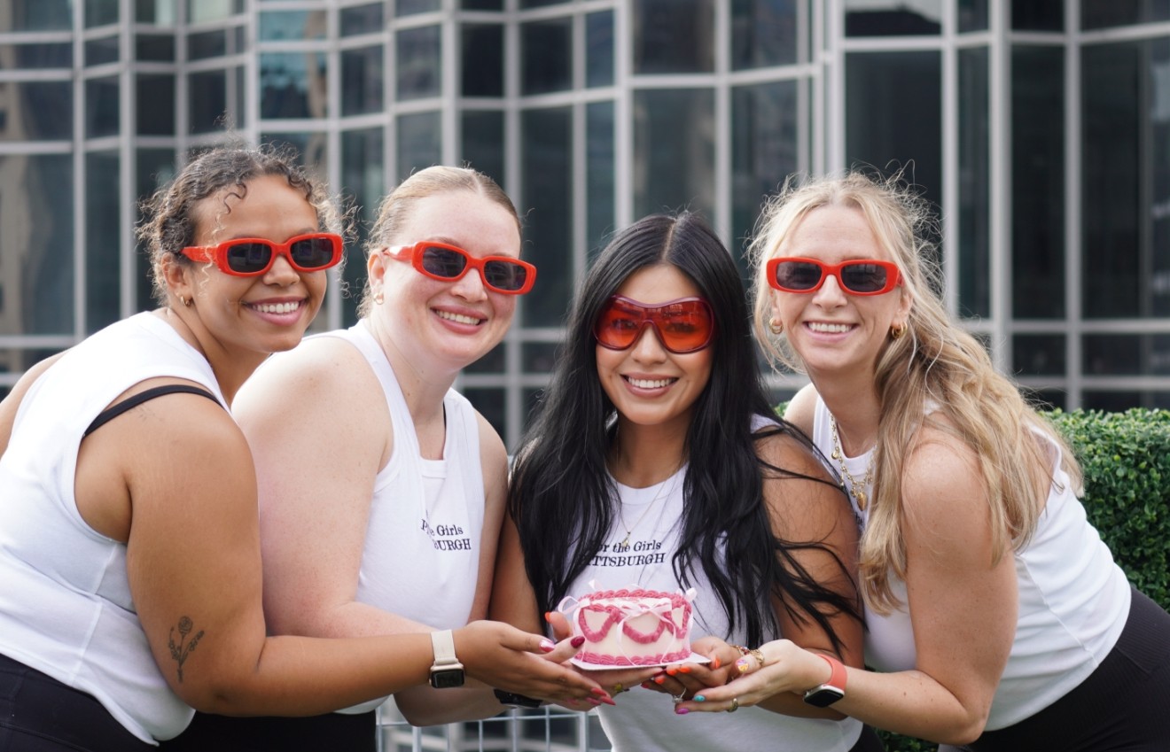 Four women wearing red sunglasses smiling outdoors, holding a cake with pink decorations, in front of a modern glass building.