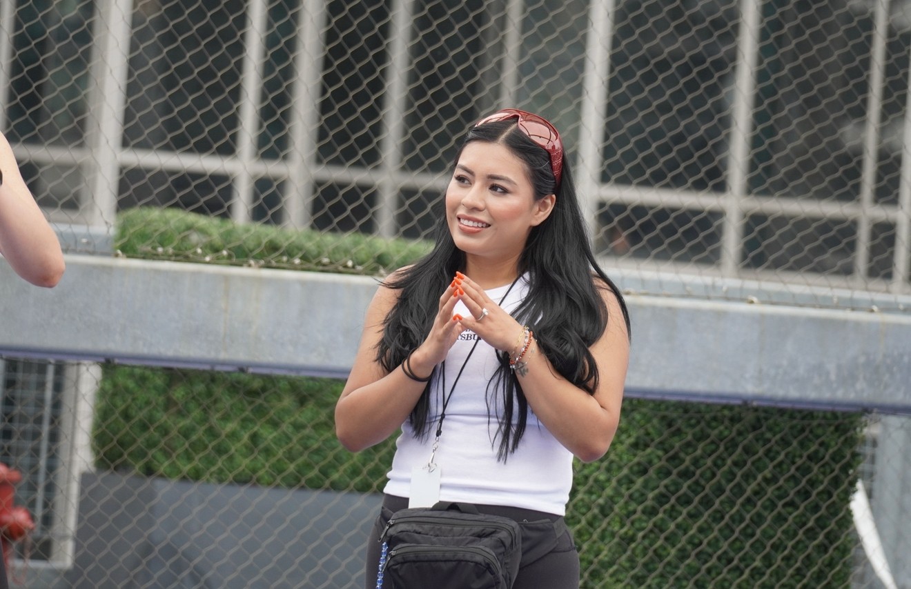 Young woman with long dark hair, smiling, standing outdoors near a chain-link fence, wearing a white top and black pants.