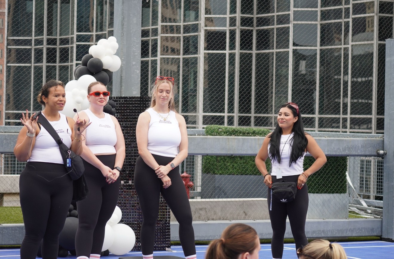 Four women stand outdoors in front of a glass building, some with hands on hips, others with arms crossed, during daytime.