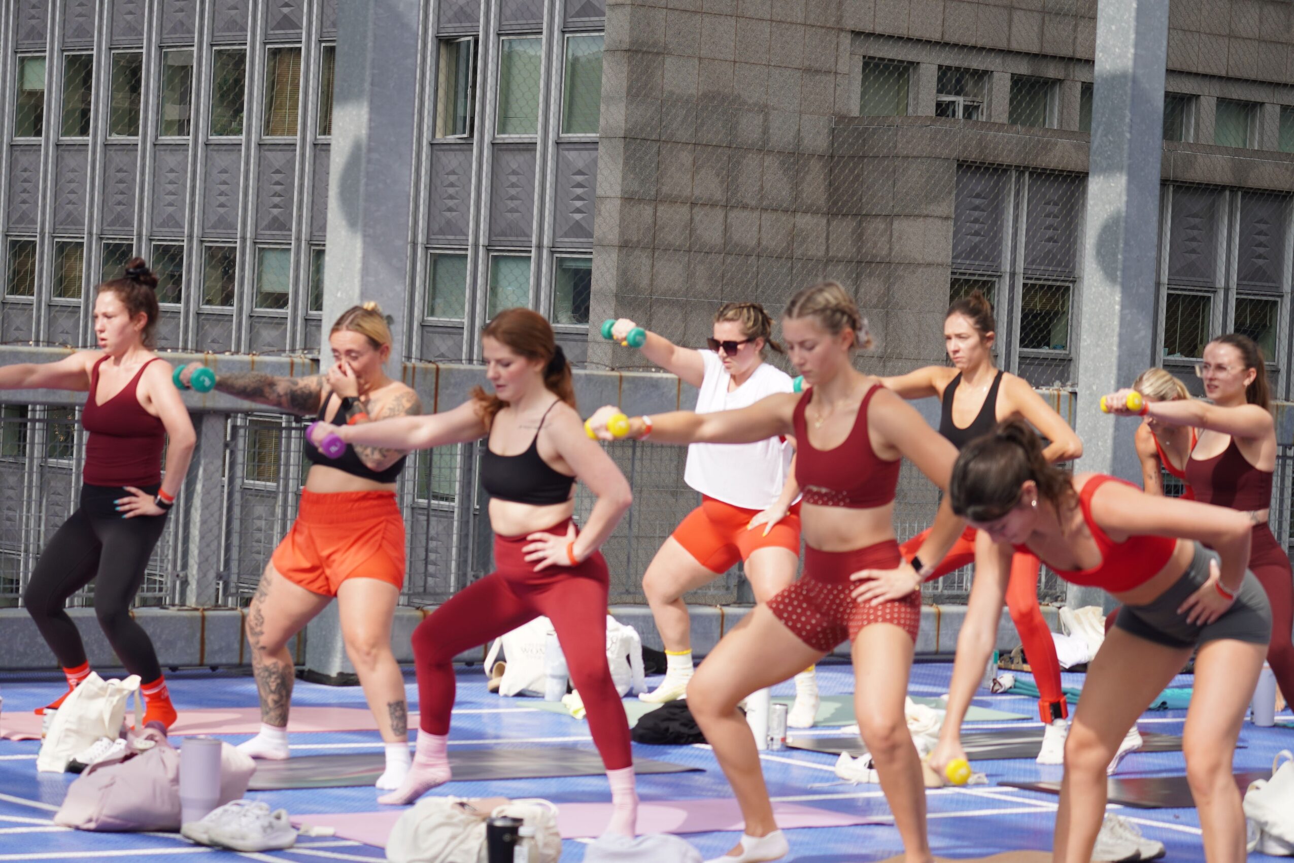 Group of women performing synchronized dance or exercise outdoors on a blue mat, with a building and metal fence in background.