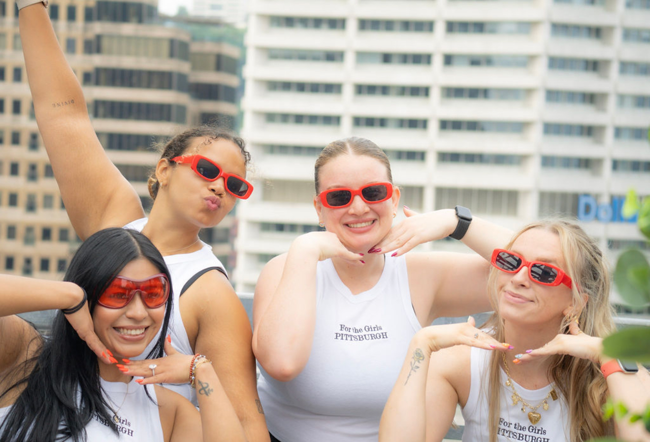 Four women wearing red sunglasses smiling outdoors with buildings in the background.