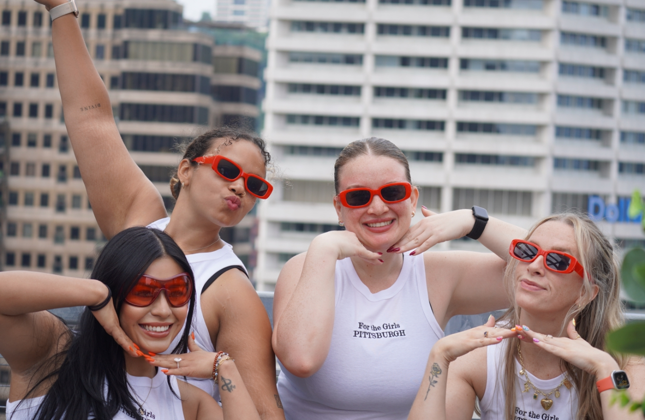 Four women wearing red sunglasses smiling outdoors with city buildings in background.