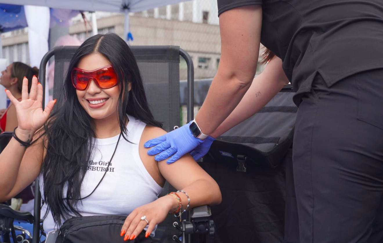 Woman with long dark hair, sunglasses, smiling, waving, receiving a vaccination from a healthcare worker outdoors.