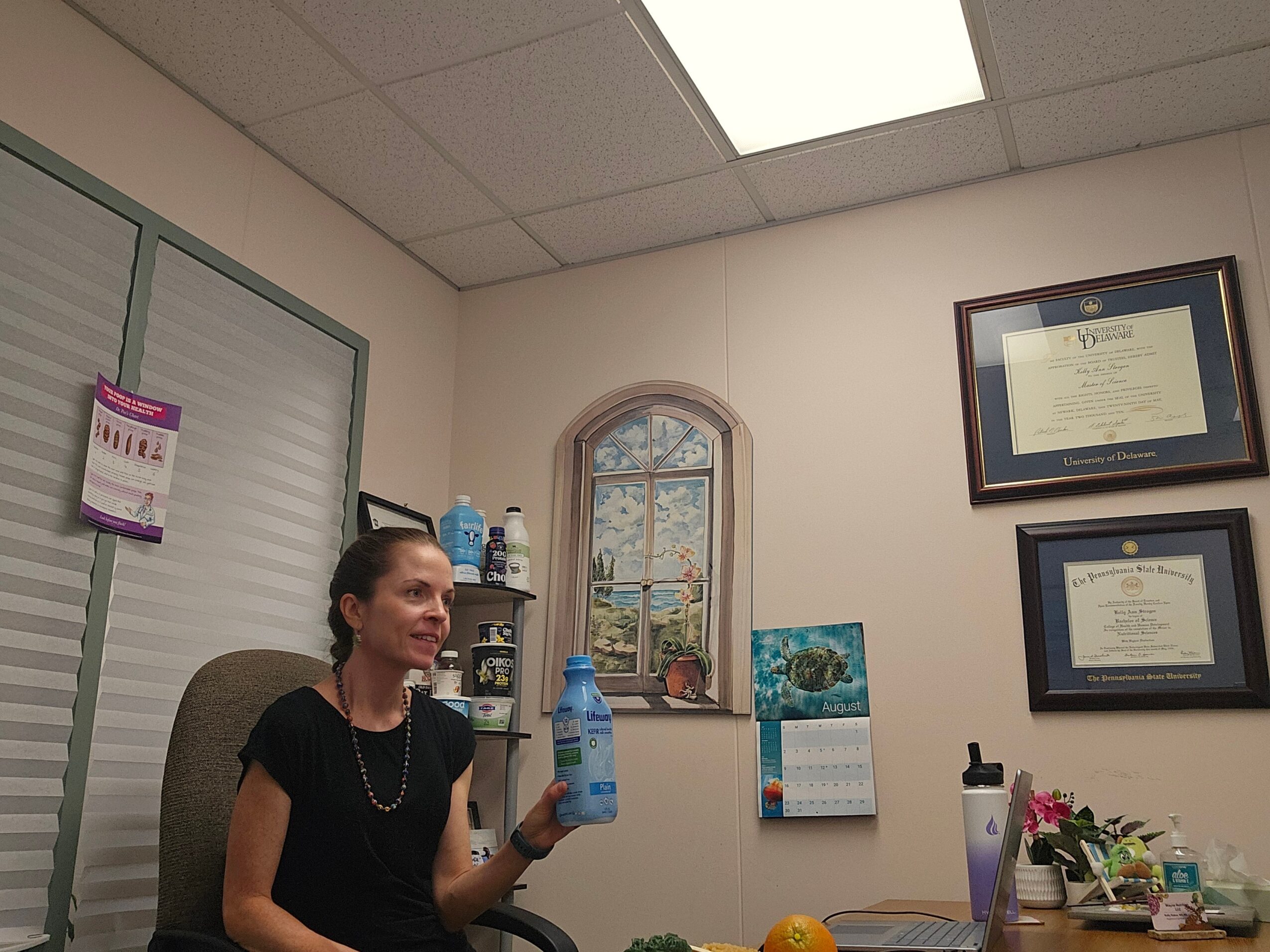 Woman sitting in an office holding a water bottle, with framed certificates on the wall and a window behind her.