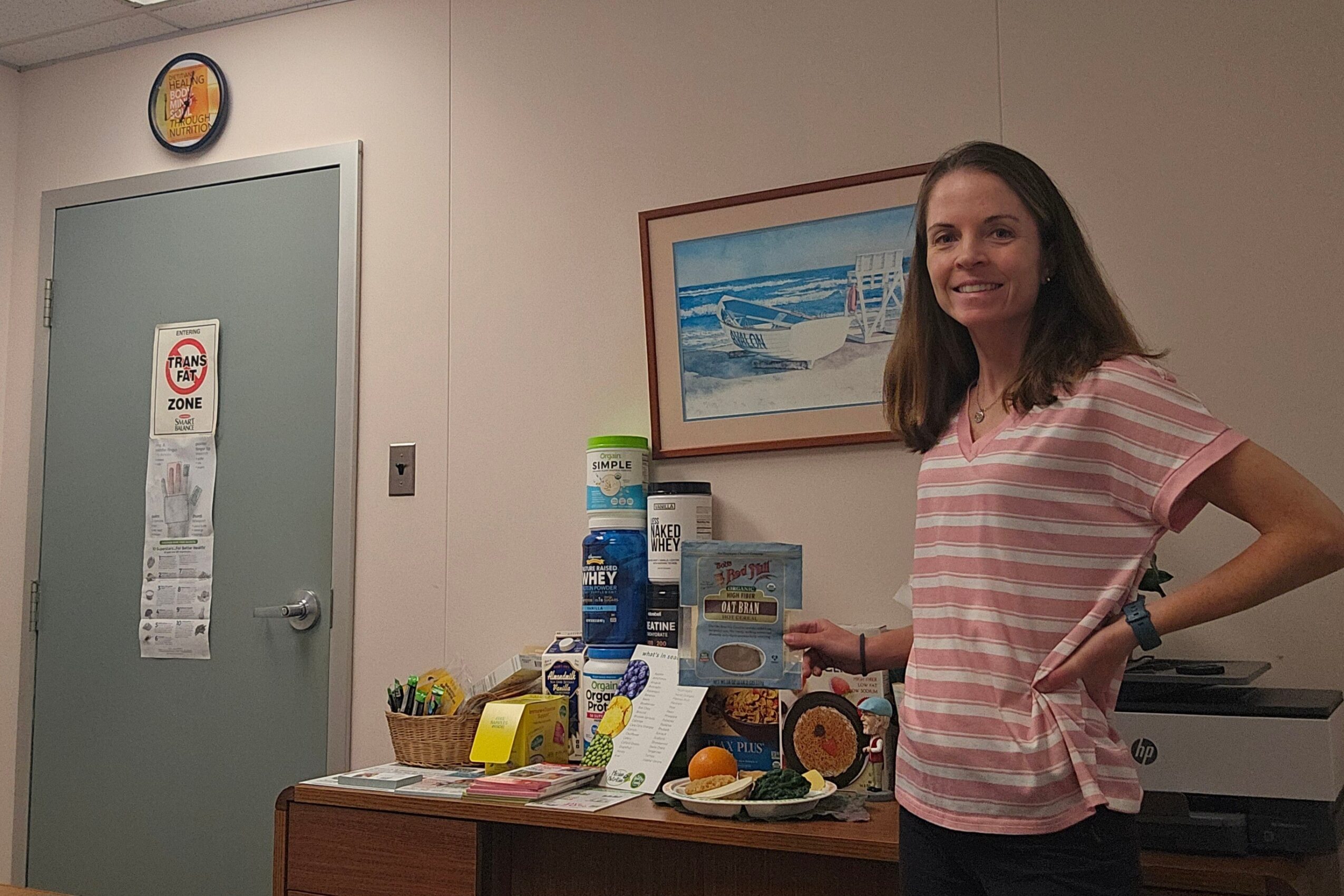 Woman standing next to a table with various items, smiling, in a room with a door and framed picture on the wall.
