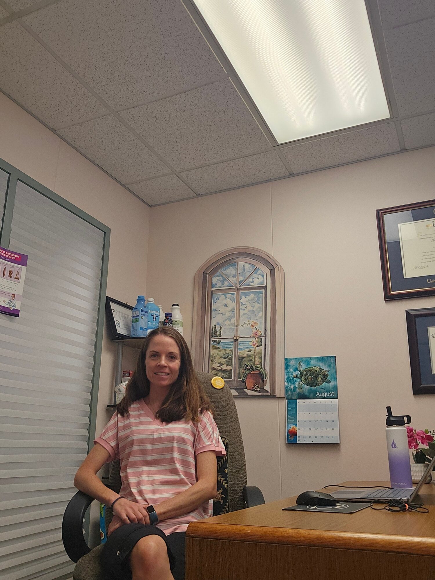 Woman standing in an office, holding a phone, with a window and calendar on the wall behind her.