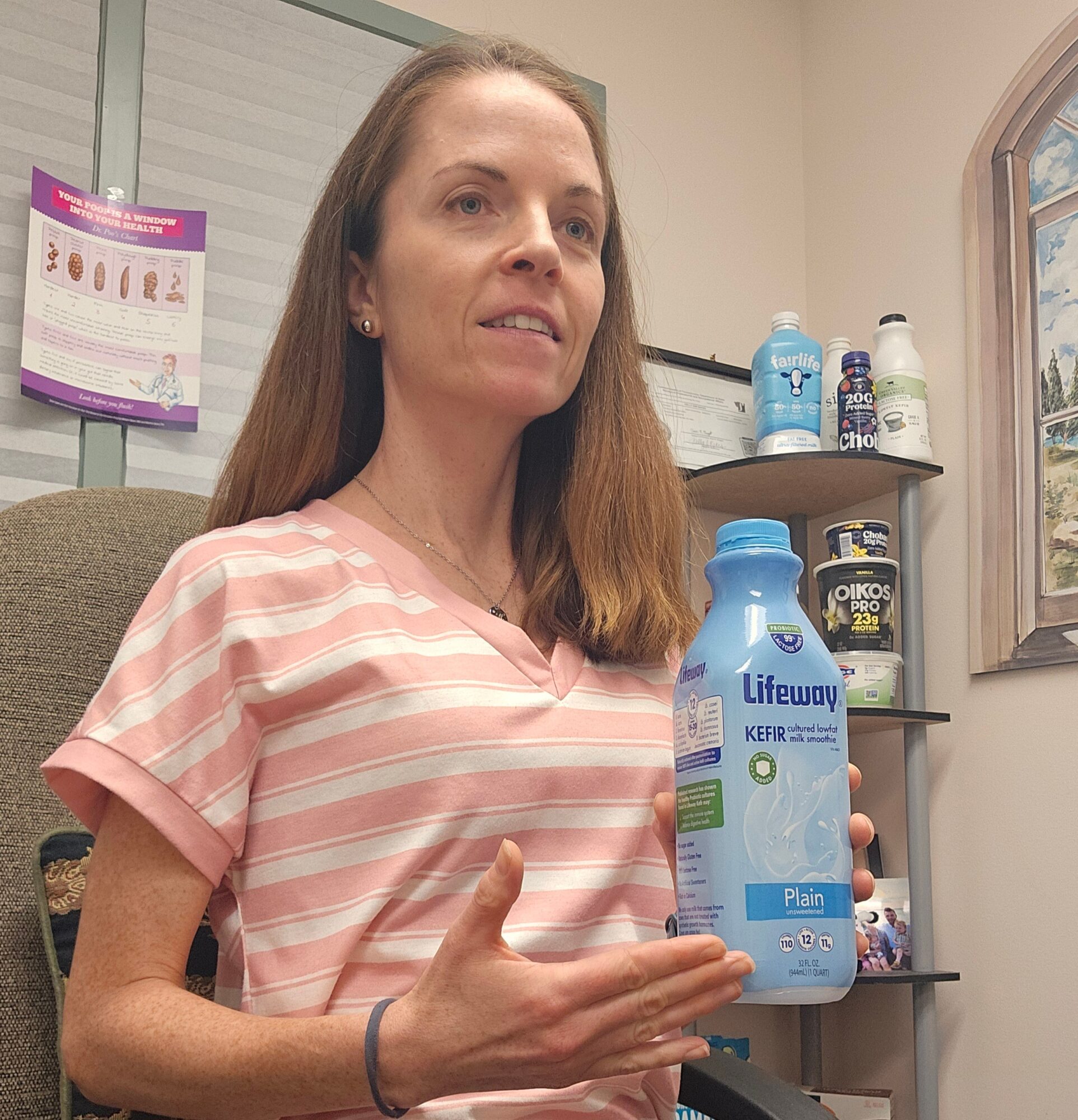 Woman with long hair in pink and white striped shirt holding a blue bottle of Lifeway kefir, smiling, indoor setting.