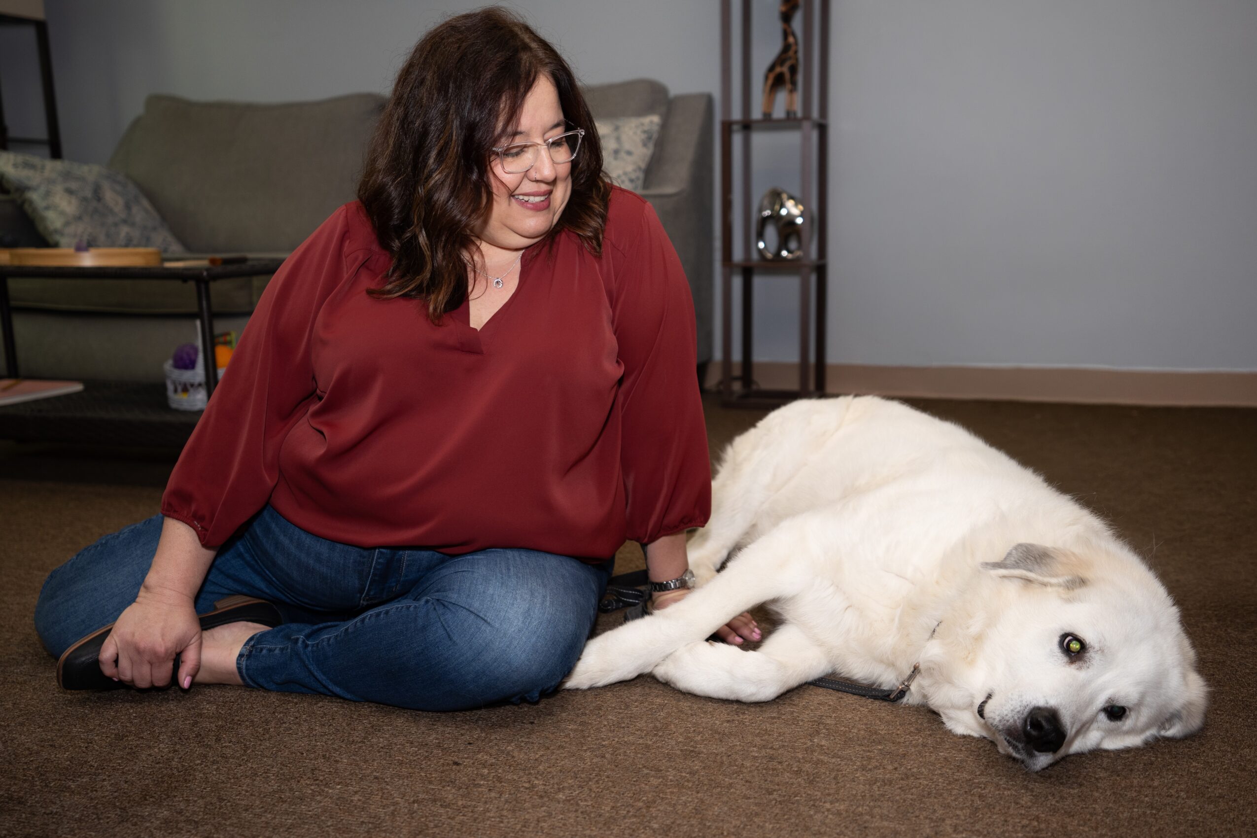 Woman sitting on the floor with a large white dog lying beside her, indoors, smiling.