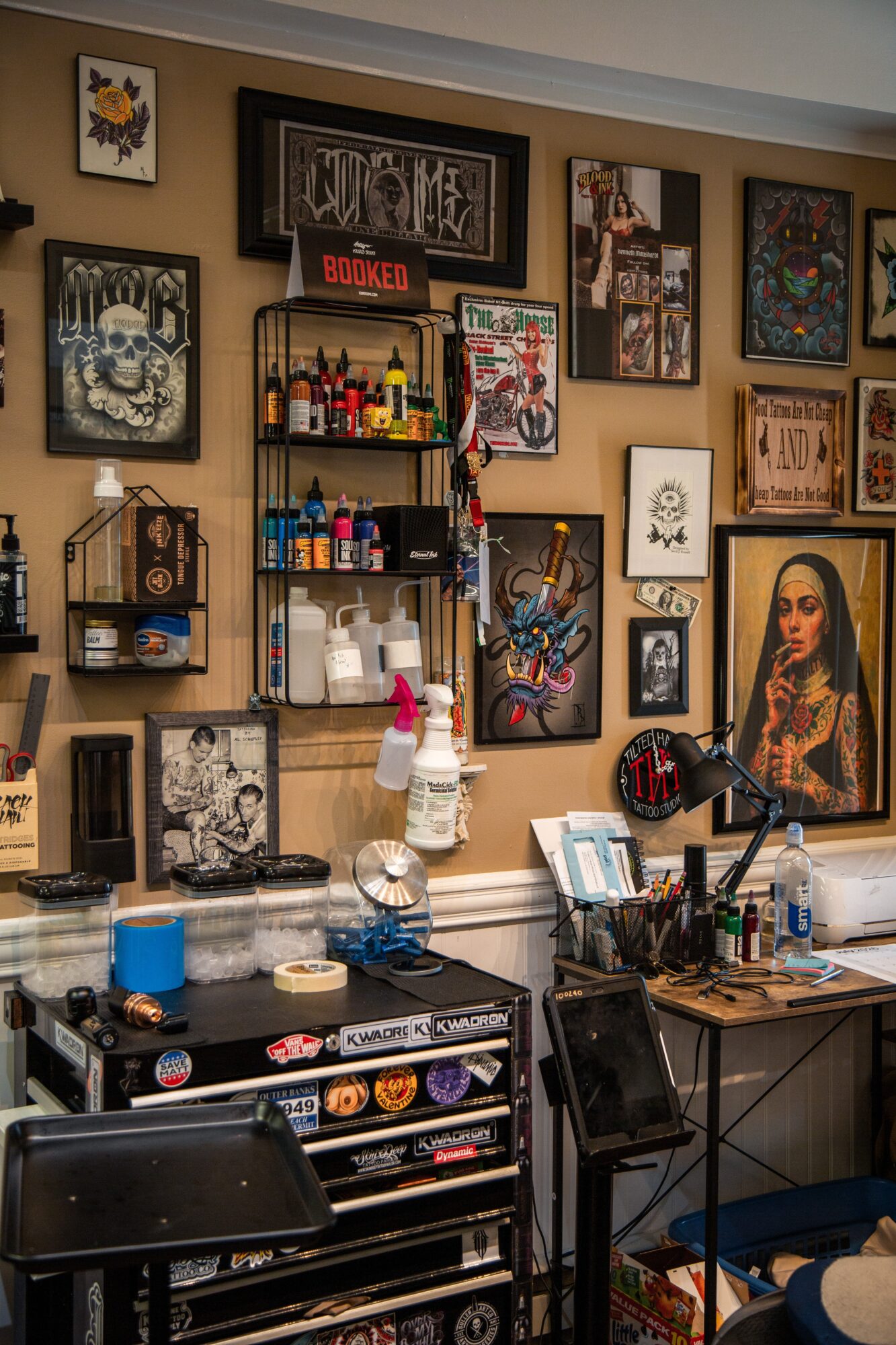 Wall decorated with framed artwork, shelves with bottles and art supplies, and a desk with art tools and a computer.