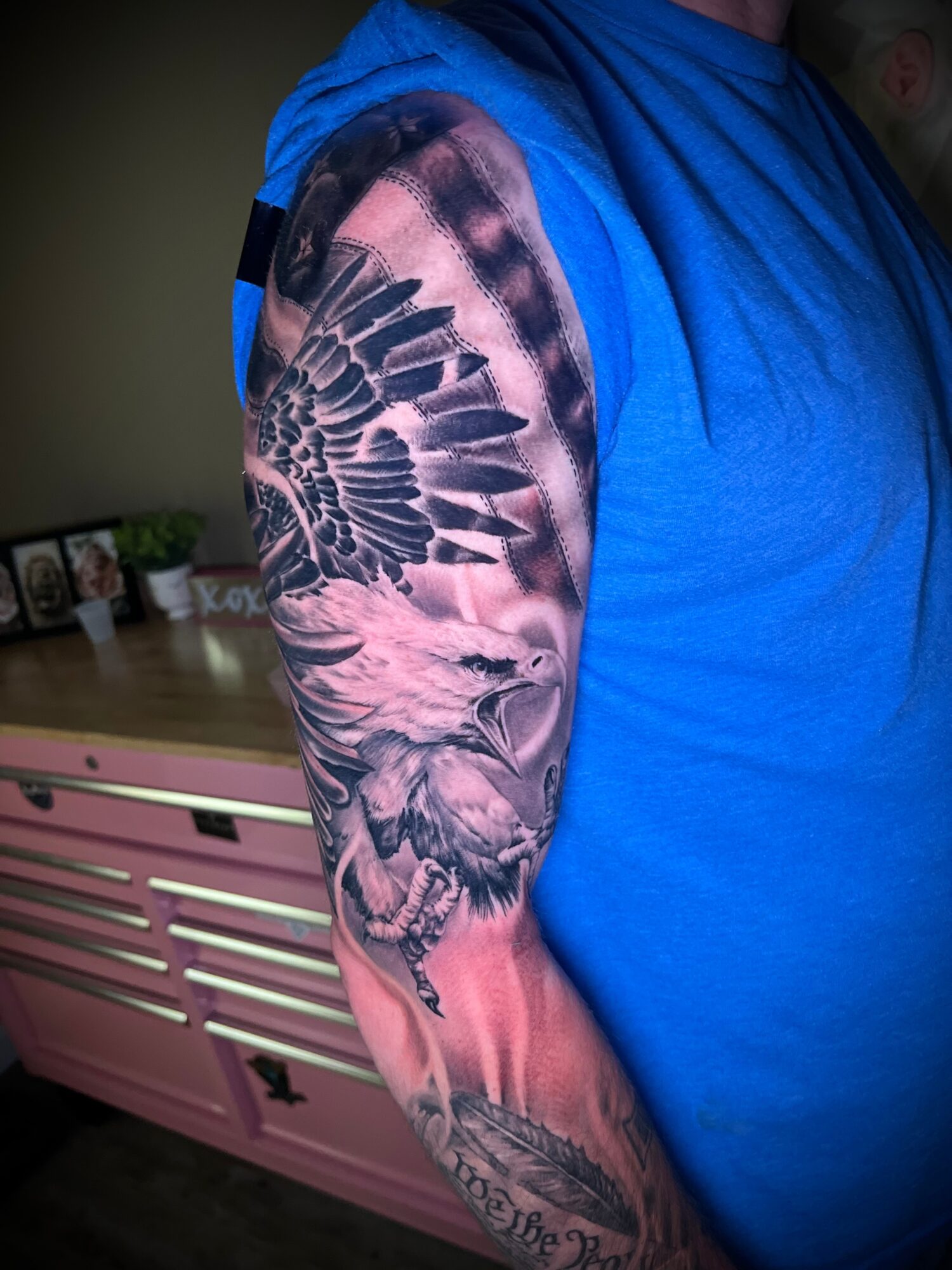 Person with a detailed black and gray skull and wing tattoo on their arm, wearing a blue shirt, indoors.