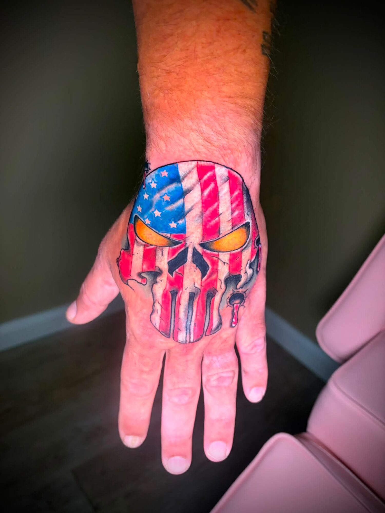 Person's hand with a colorful skull tattoo featuring the American flag design, visible on the back of the hand.