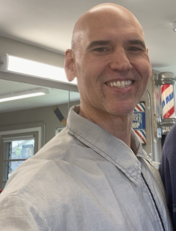 Smiling bald man in a light-colored shirt indoors, with barber poles in the background.