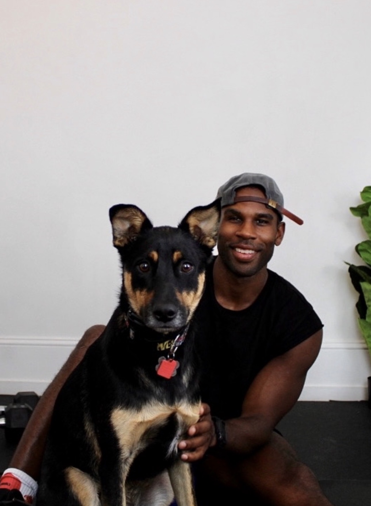 Man smiling with a black and tan dog indoors, plain white wall background, green plant on the right.