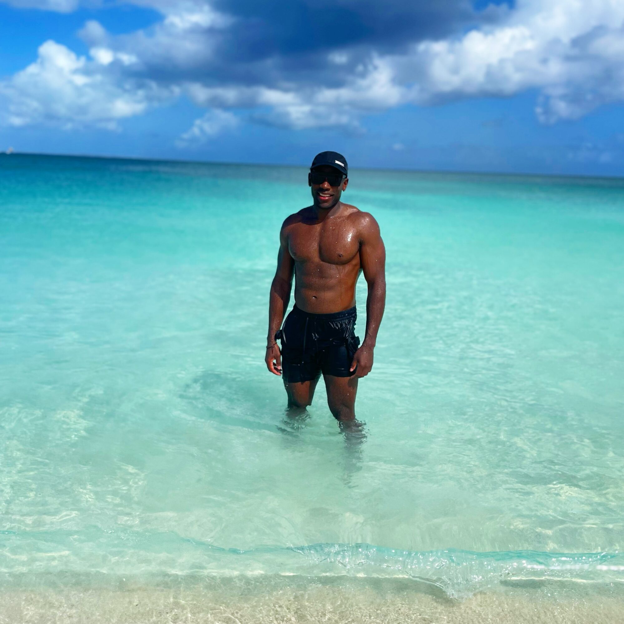 Man standing in clear turquoise ocean water, wearing sunglasses, black shorts, with a partly cloudy sky in the background.