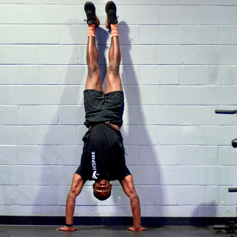 Person performing a handstand against a wall, legs extended upward, wearing black shorts and a black T-shirt, in a gym setting.