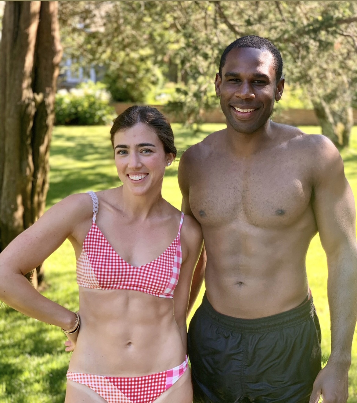 Two people smiling outdoors, one woman in a red checkered bikini top and one shirtless man, standing on grass with trees in background.