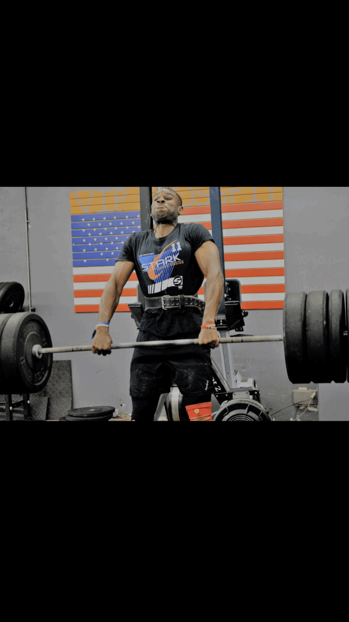 Person lifting a heavy barbell in a gym with an American flag in the background.
