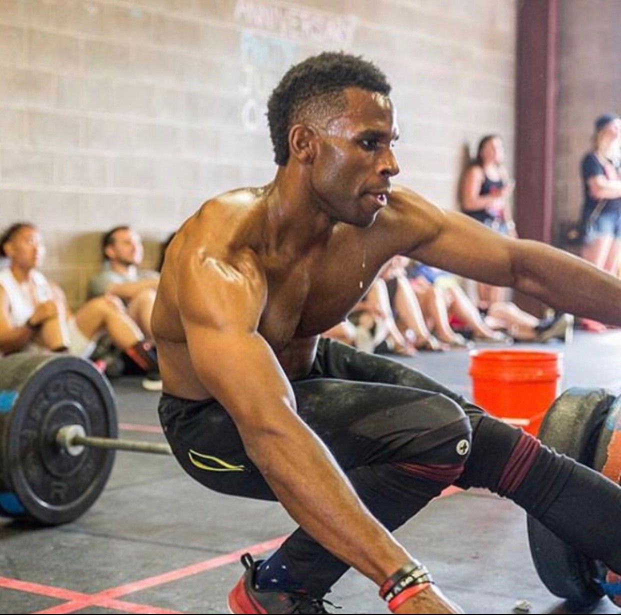 Man exercising with a barbell on the floor, surrounded by people in a gym, some sitting and some standing, in a spacious room.