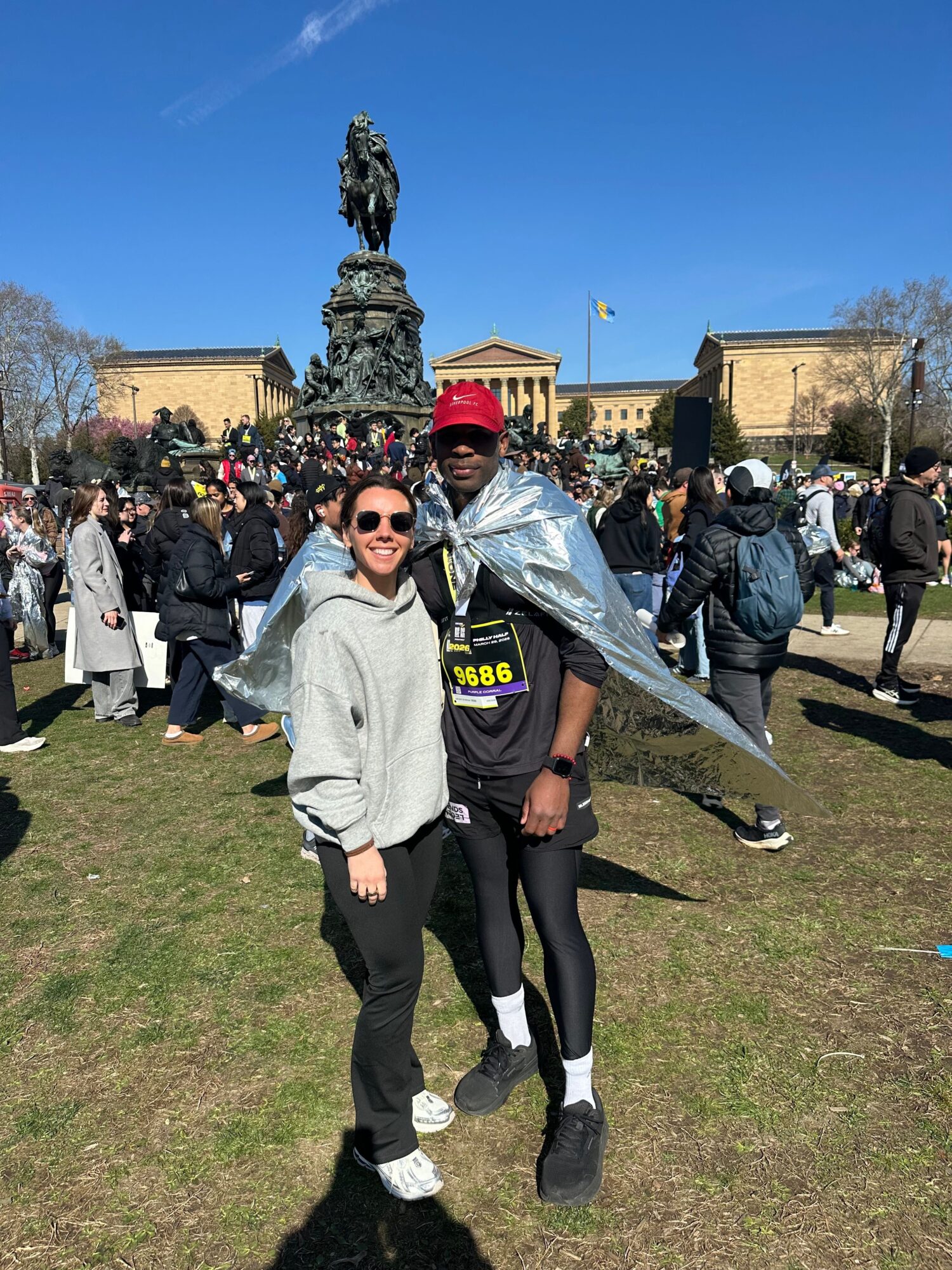 Two women standing on grass, one wearing sunglasses and a gray jacket, the other in a black jacket with a red headscarf, with a crowd and structures in the background.