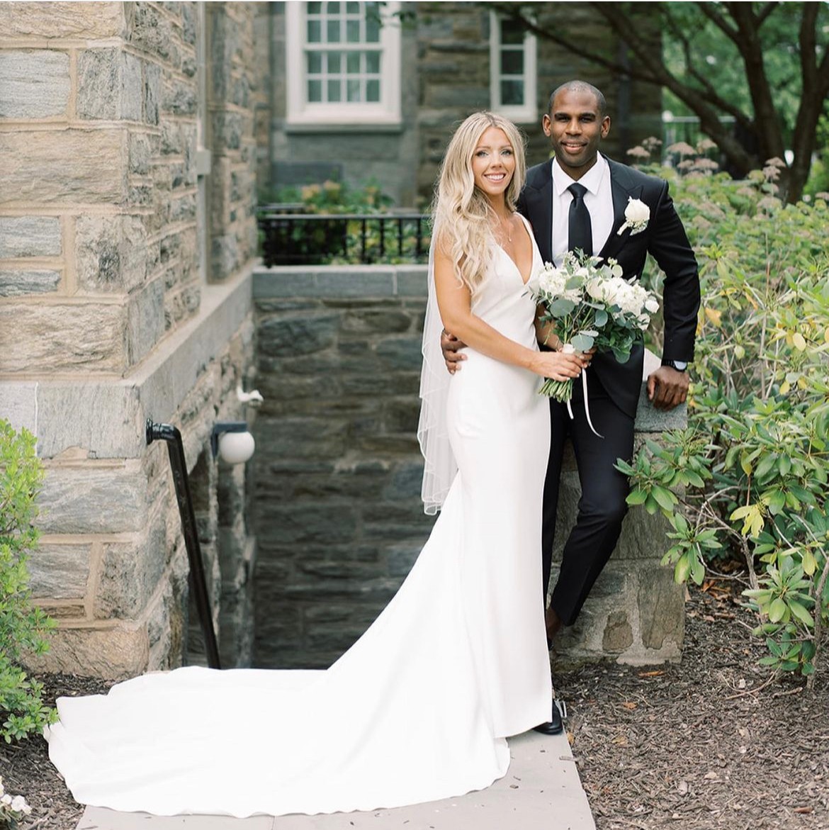 Bride and groom standing outdoors, smiling, with greenery and stone building in background, bride holding bouquet, groom in suit.