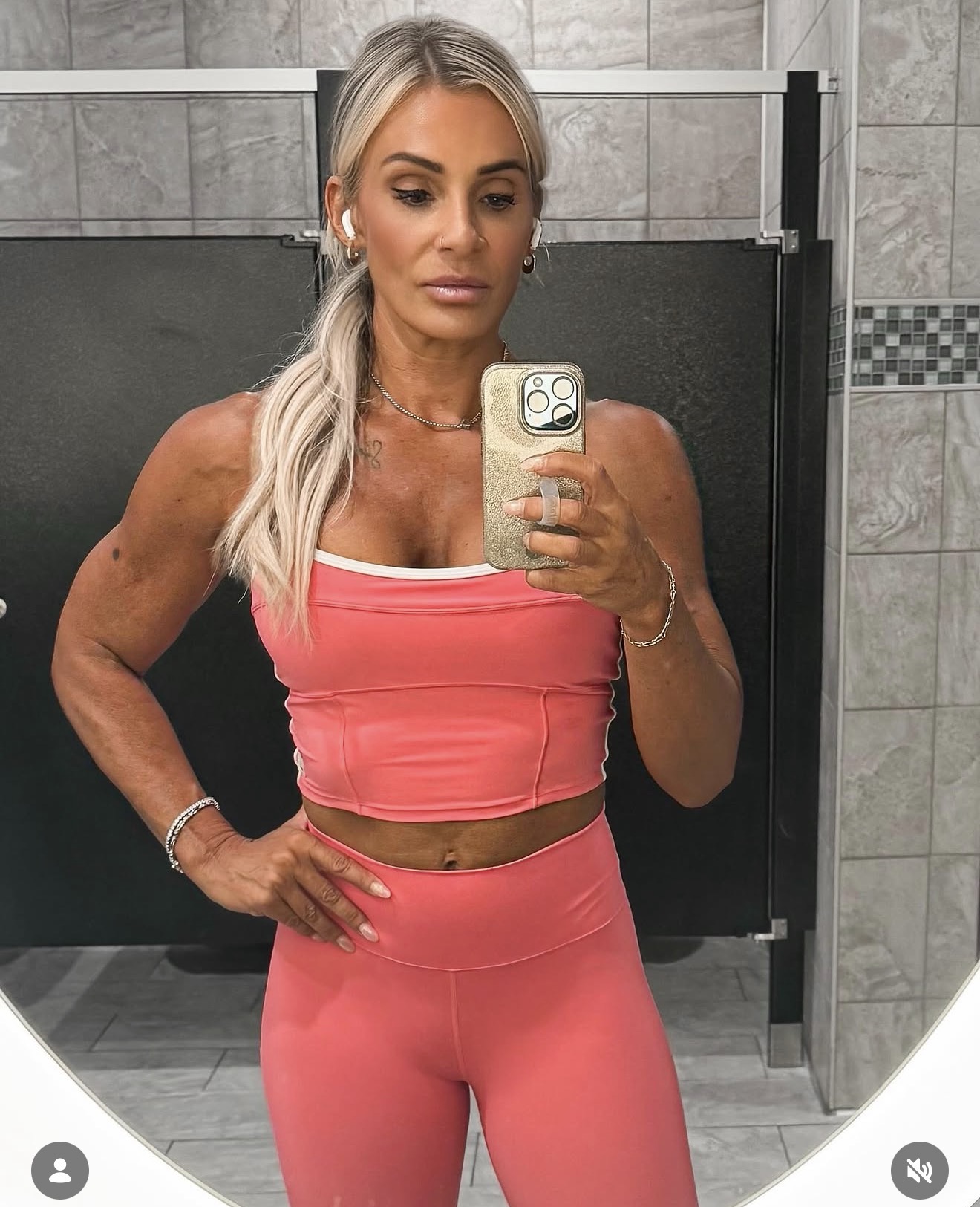 Woman with long blonde hair taking mirror selfie in a bathroom, wearing a pink sports bra and matching leggings.