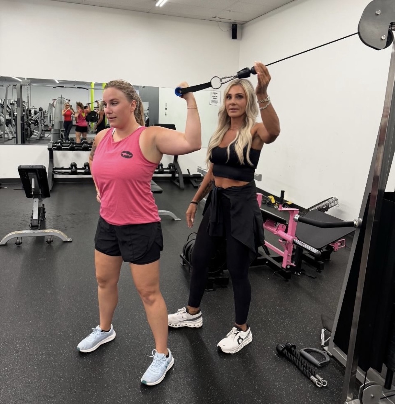 Two women in workout clothes using a barbell with a weight plate in a gym, one lifting, the other spotting, mirrors in background.