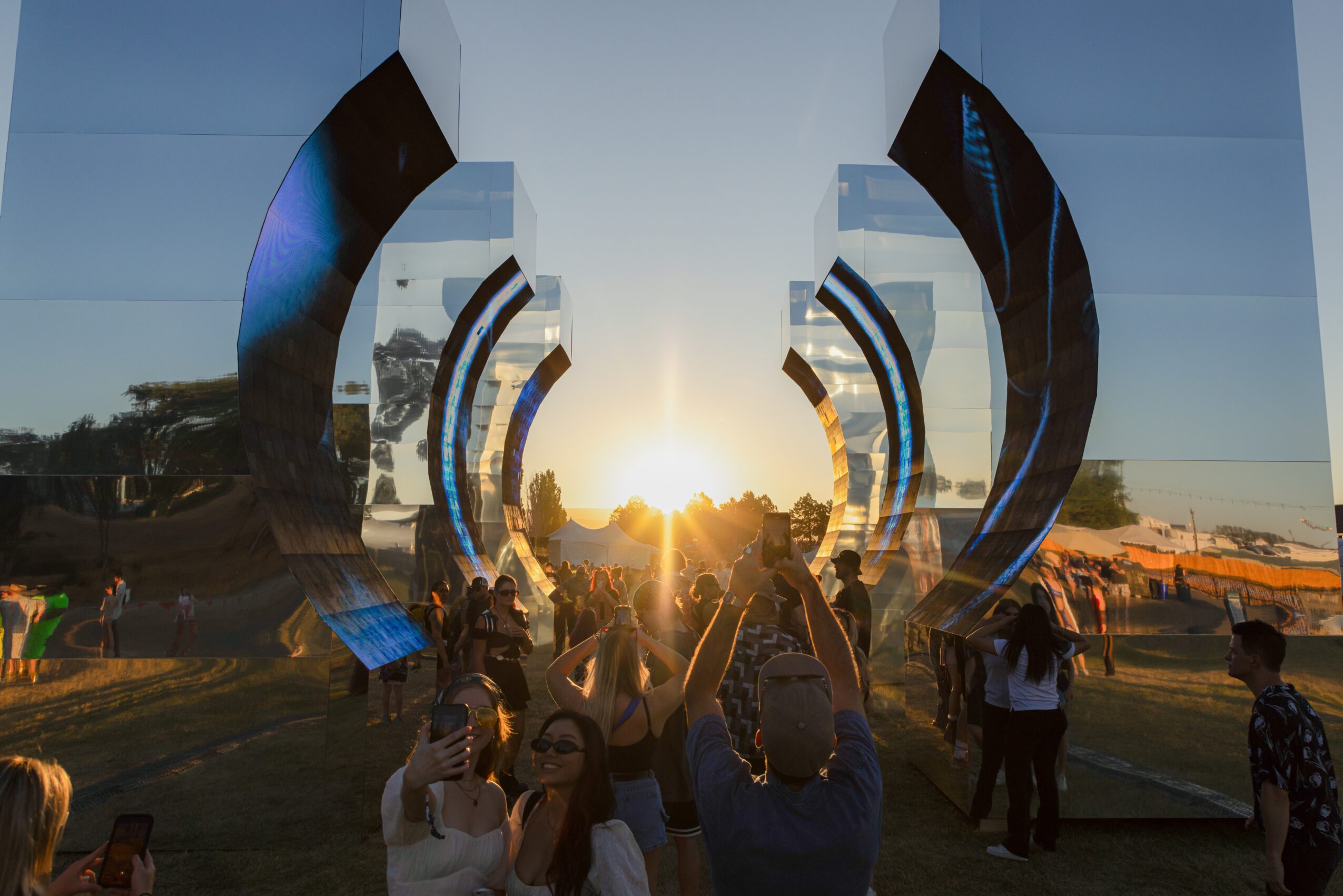 Sunset scene with people gathered near reflective, curved structures outdoors, with a bright sun on the horizon.