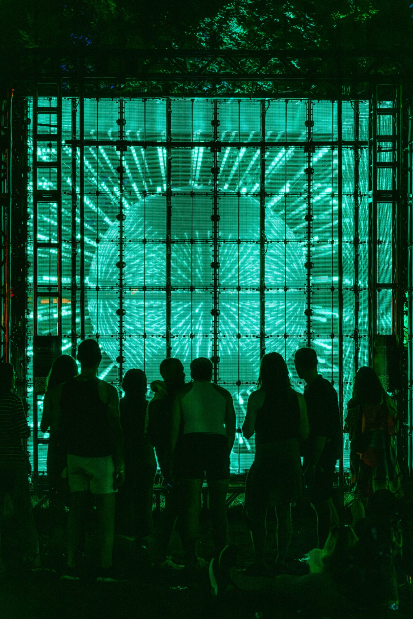 Group of people standing in front of a large, illuminated, teal-colored grid structure at night.