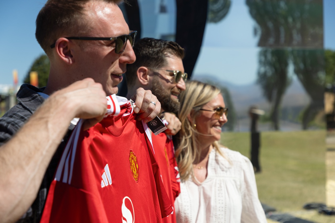 Three people smiling outdoors, one holding a red sports jersey, with trees and a fence in the background.