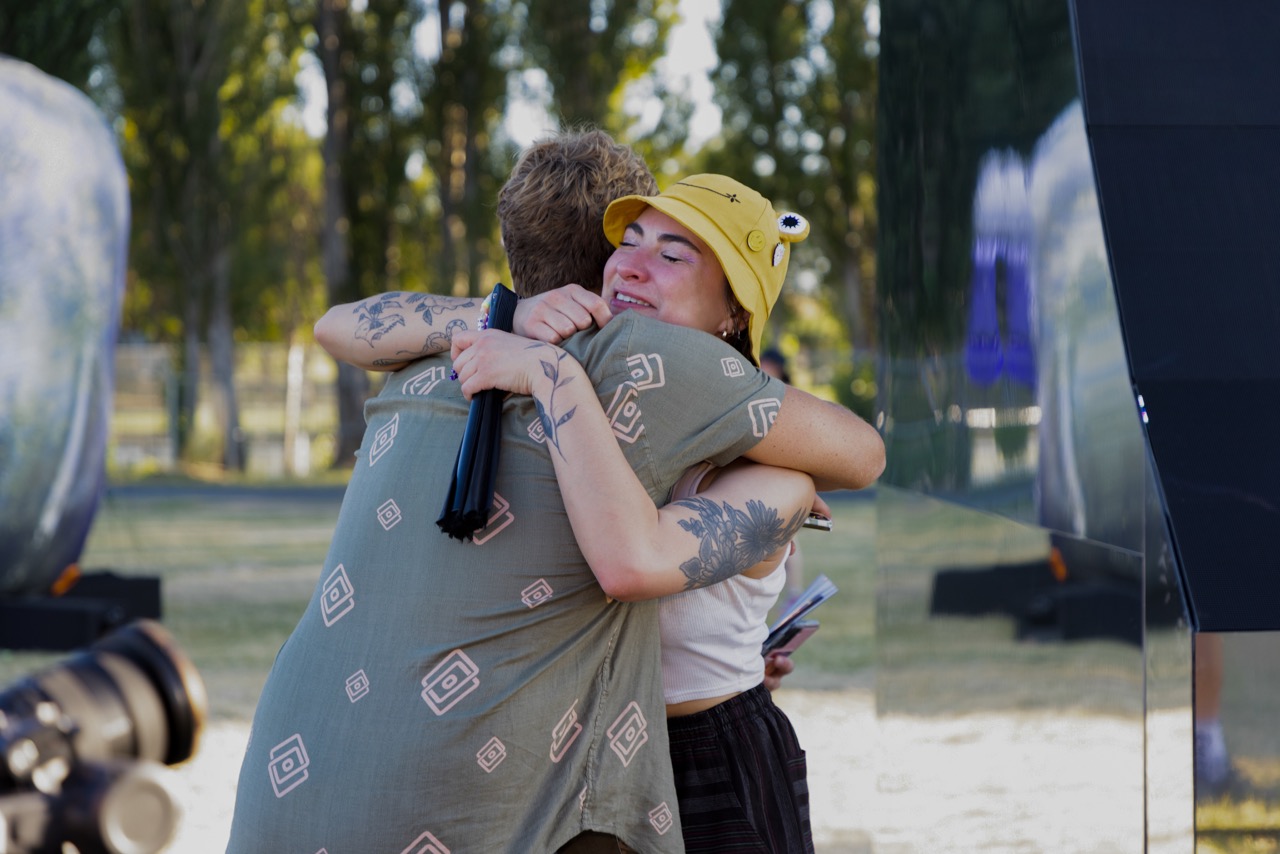 Two people hugging outdoors, one wearing a yellow hat, trees in the background, and a reflective surface nearby.