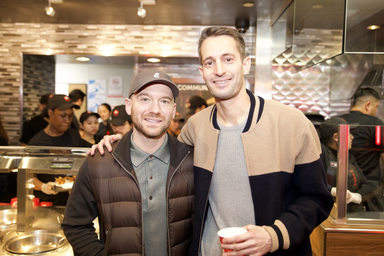 Two men smiling, one with arm around the other, in a busy restaurant or cafe setting.