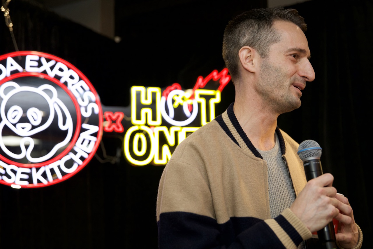 Man holding microphone on stage with neon signs in background, including a panda logo and the words 'HOT ONE'.